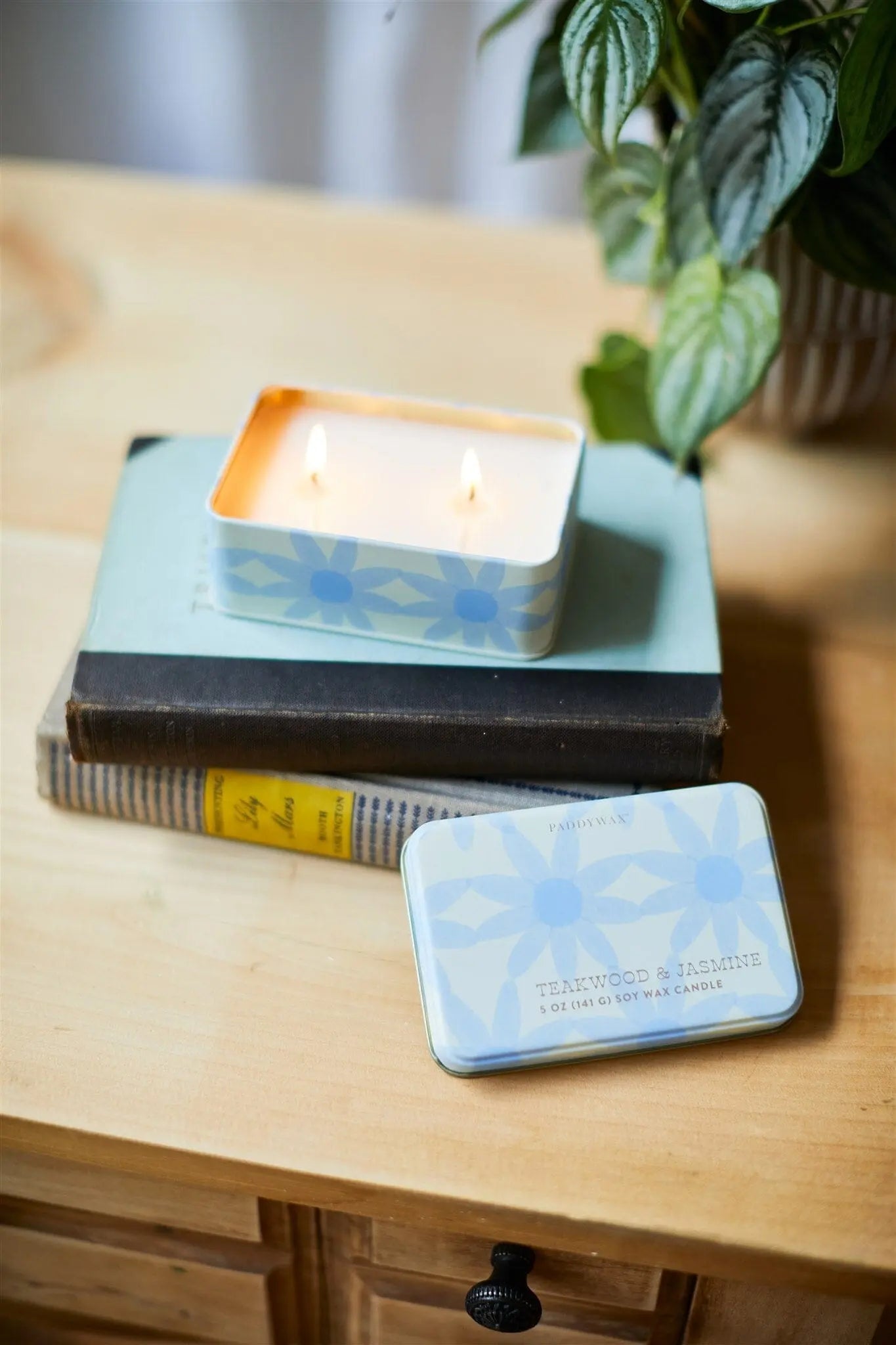 An Everyday Tins 5 oz. Candle in Teakwood & Jasmine sits lit on books atop a wooden table, its lid nearby. The sweet fragrance fills the air as a potted plant brings extra charm to the scene.