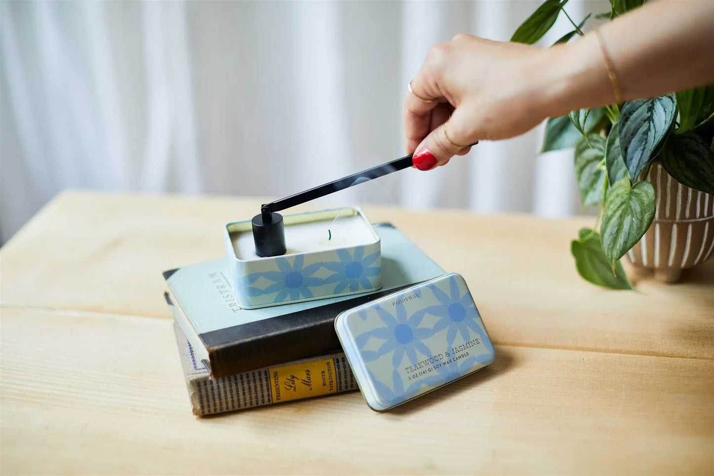 A hand with red nail polish uses the Black Candle Snuffer to extinguish candles in a blue floral tin, set on stacked books beside a potted plant on a wooden surface.