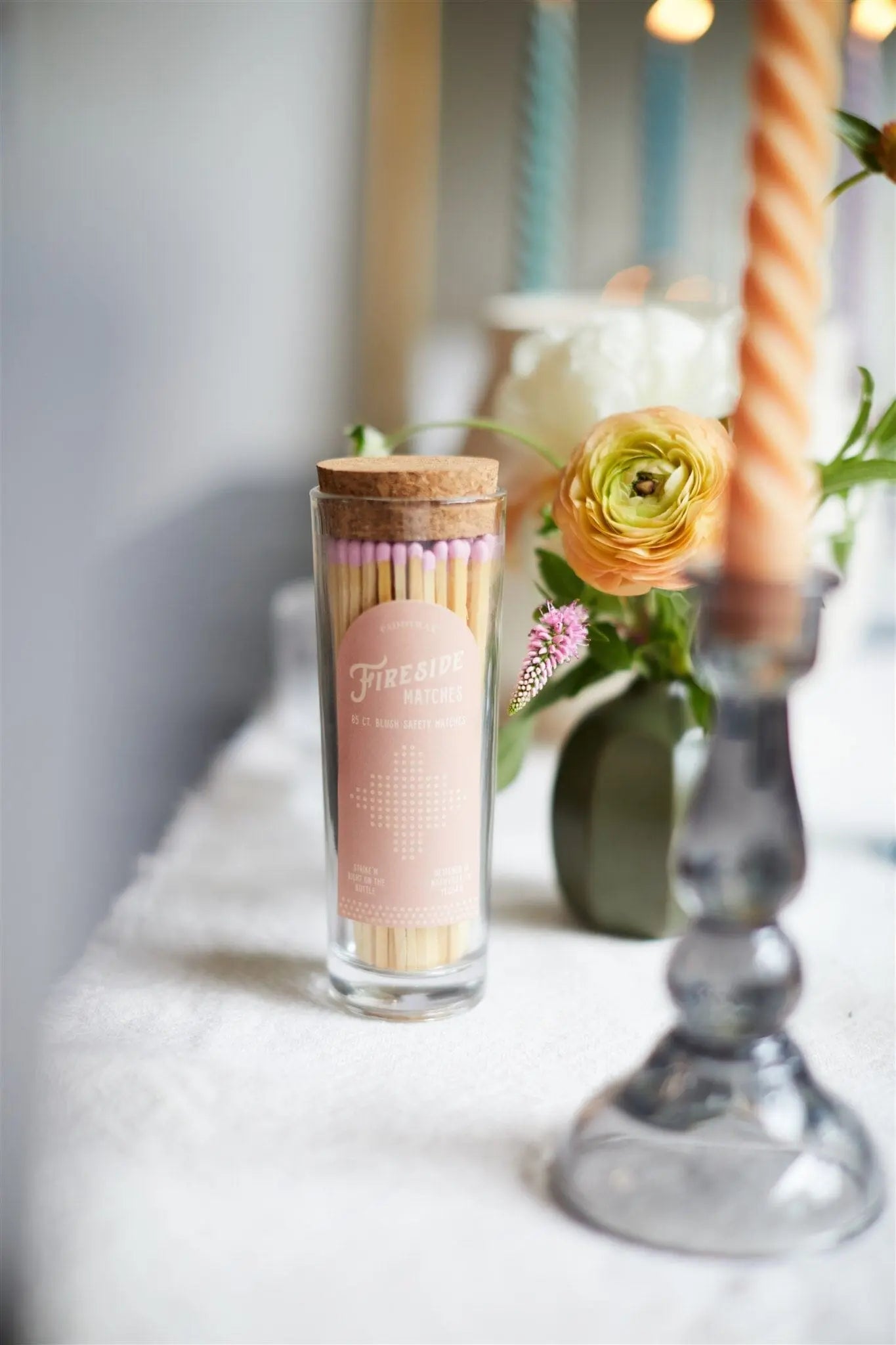 A glass jar of Fireside Safety Matches - Blush, featuring pink-tipped matches for a vintage vibe, sits on a white tablecloth beside a flower arrangement and a twisted candle in a holder.