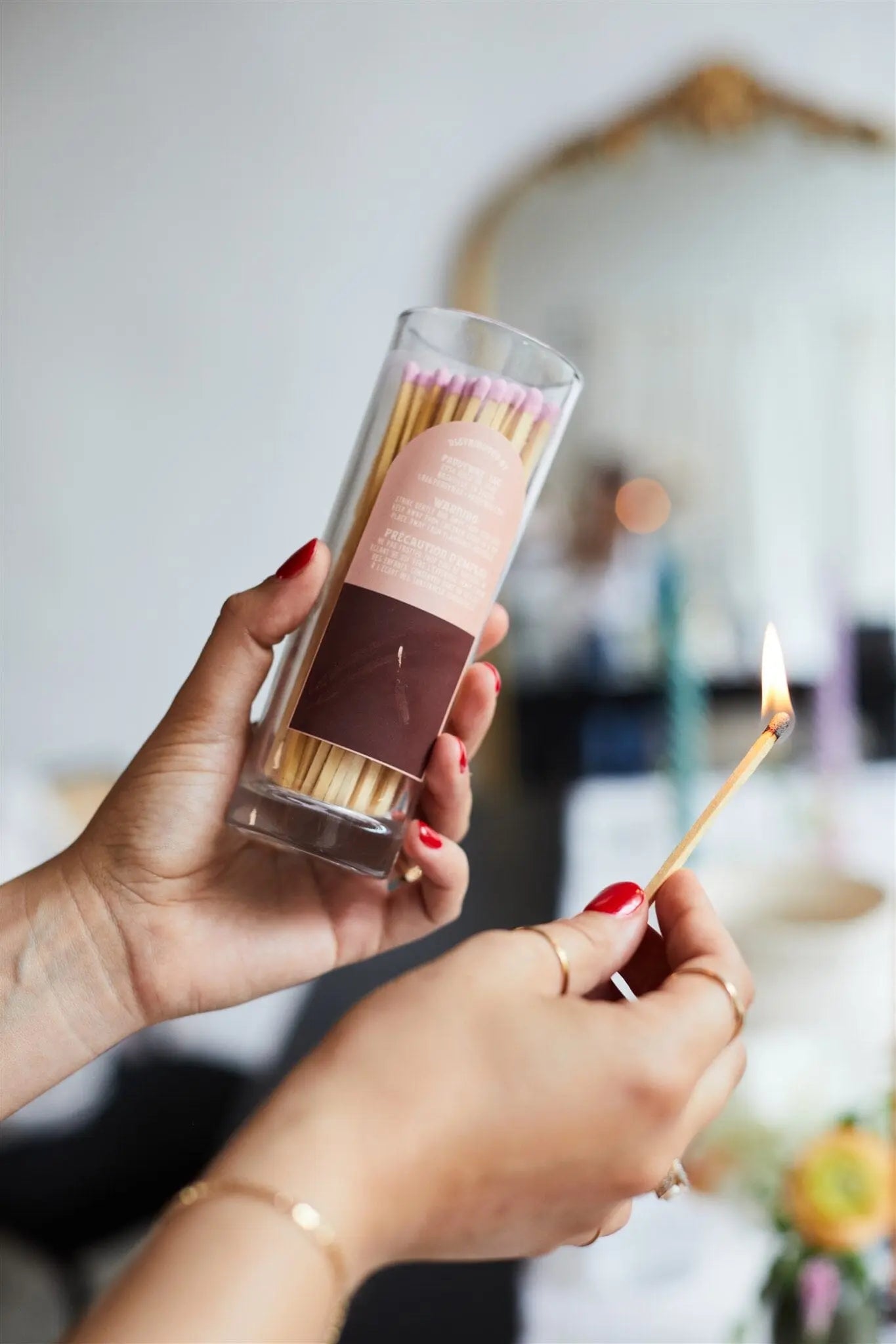 A person holds a clear jar of Fireside Safety Matches - Blush in one hand and lights a match with the other, against a softly blurred background that adds a vintage touch.
