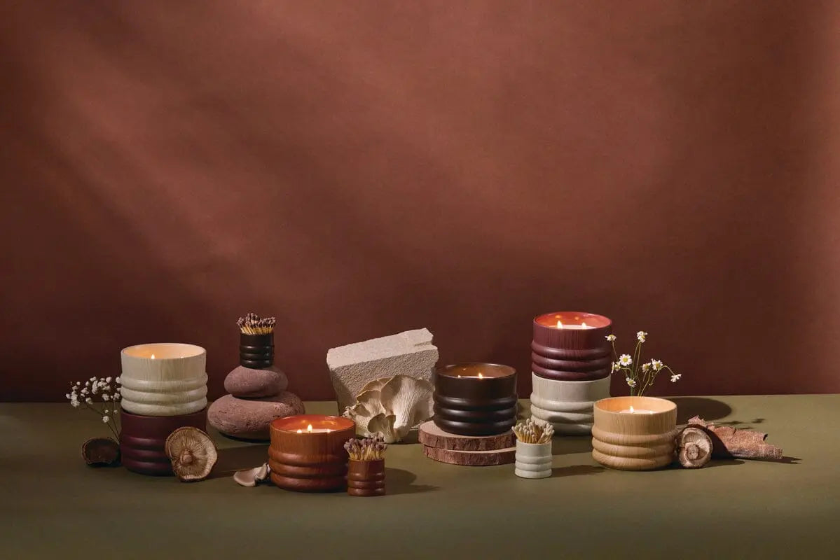 Five Grain Candle - Bergamot & Mahogany ribbed ceramic candles in earth-toned holders are arranged with small match containers, dried flowers, stones, and woodgrain-inspired slices on a green surface against a brown background.