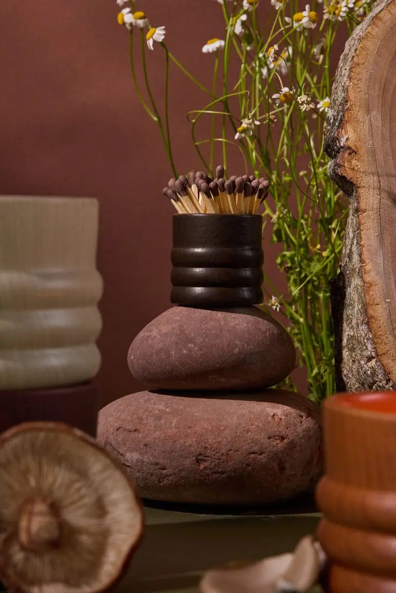 A stack of smooth stones topped with the Dark Brown Ceramic Wood Match Holder, surrounded by flowers, a tree slice with visible woodgrain, and textured ceramic vessels.