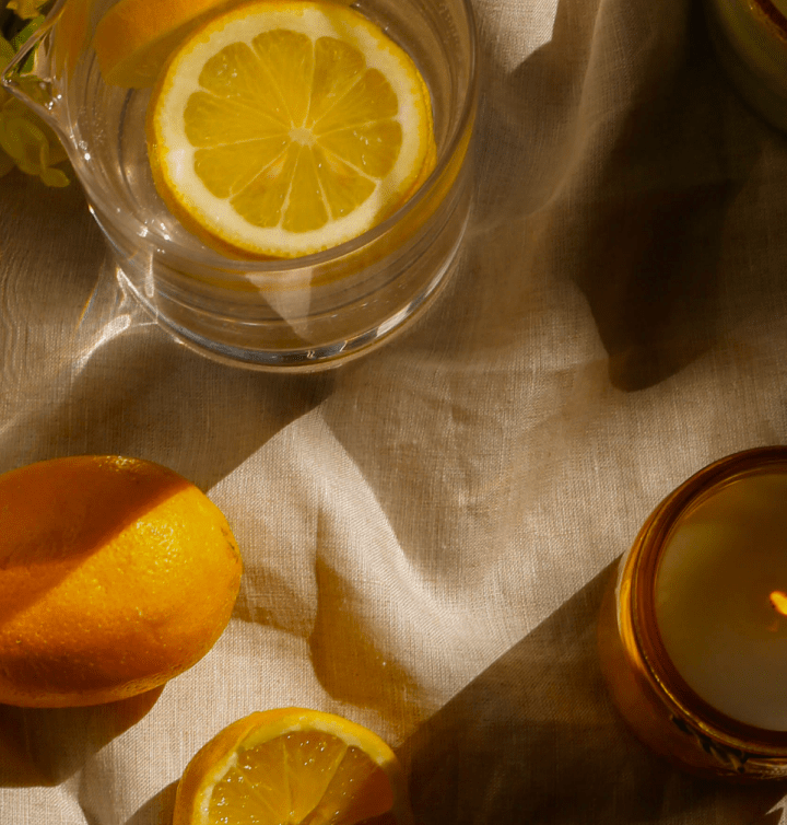 A glass of water with a lemon slice, a whole lemon, a half lemon, and a lit candle arranged on a beige fabric surface in natural sunlight.