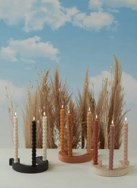 Three spiral taper candles in colorful holders sit before dried grass, with a blue sky backdrop. The Nashville Black Ceramic Horseshoe Taper Holder w/ Hangtag adds a unique touch to this charming home decor scene.
