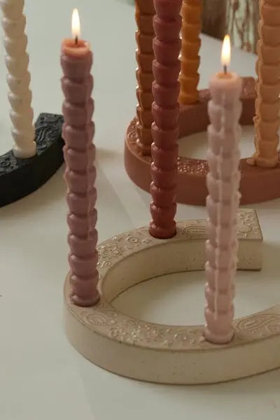 Several tall, spiral candles in assorted colors are lit and placed in curved, textured holders, including the Nashville White Ceramic Horseshoe Taper Holder w/ Hangtag, adding a stylish touch to your home decor.