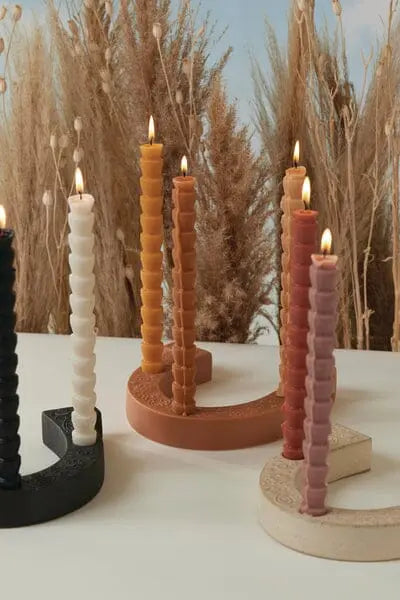 Five lit spiral candles in various colors stand in Nashville Terracotta Ceramic Horseshoe Taper Holders on a white surface, with tall dried grasses in the background.