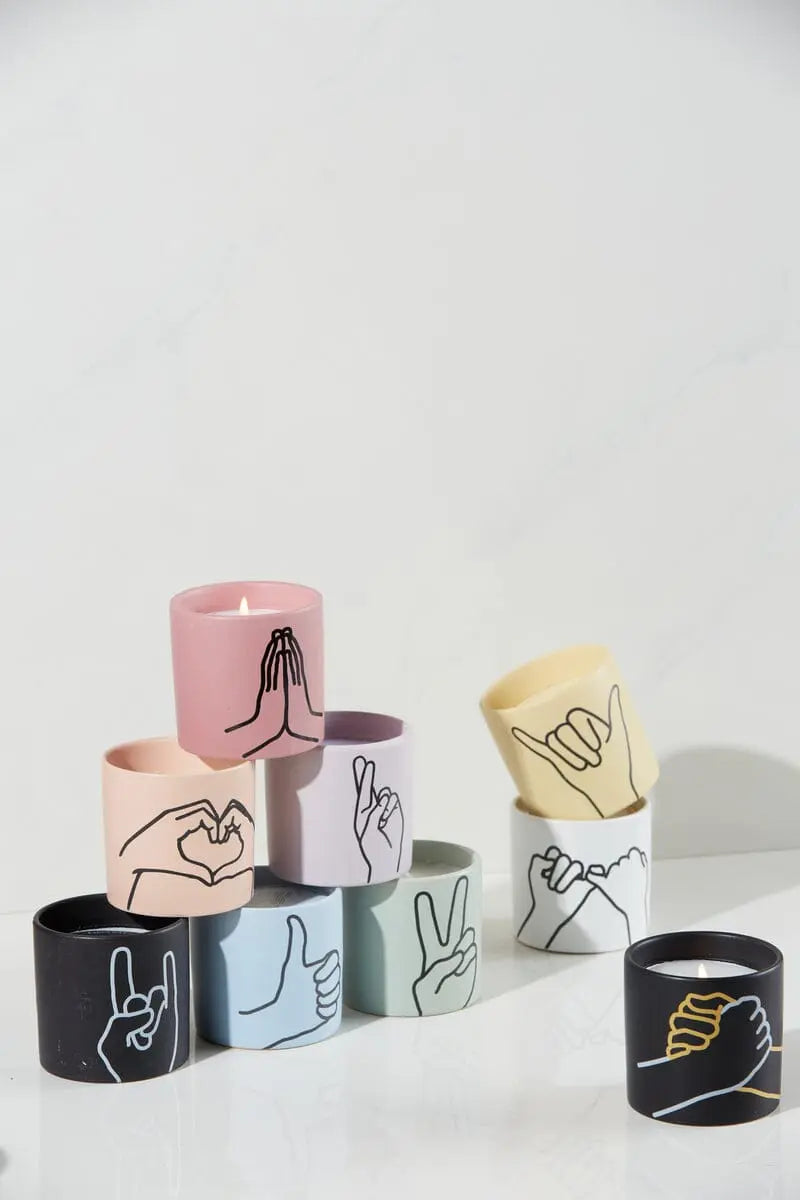 The Impressions 5.75 oz Candle - Leather + Oakmoss Rock On features a colorful matte ceramic vessel with a hand gesture illustration in black or yellow line art and gold foil stamped details for a unique, charming look.