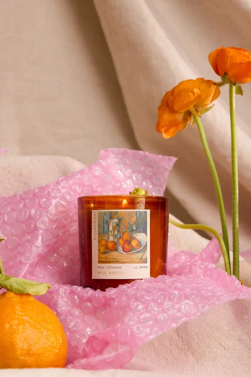 The Impressionist Candle - Cezanne Wild Neroli, an orange art candle in a glass jar with a Cézanne label, rests on pink bubble wrap beside an orange flower and tangerine, set against a beige fabric background.