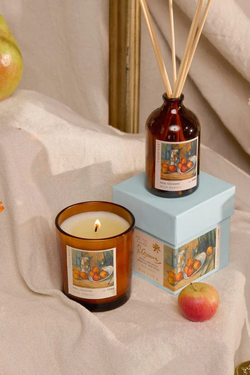 The Impressionist Diffuser- Cezanne Wild Neroli, with its reed sticks and elegant blue box, sits on beige fabric alongside apples and a gold frame, creating a warm, Impressionist-inspired ambiance.