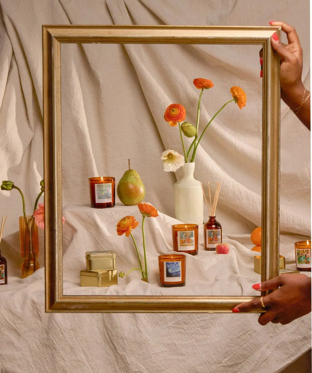 A person holds a gold frame around the Impressionist Candle – Claude Monet Secret Garden, surrounded by candles, flowers, and a pear on draped beige fabric, evoking the soft beauty of Impressionist art.