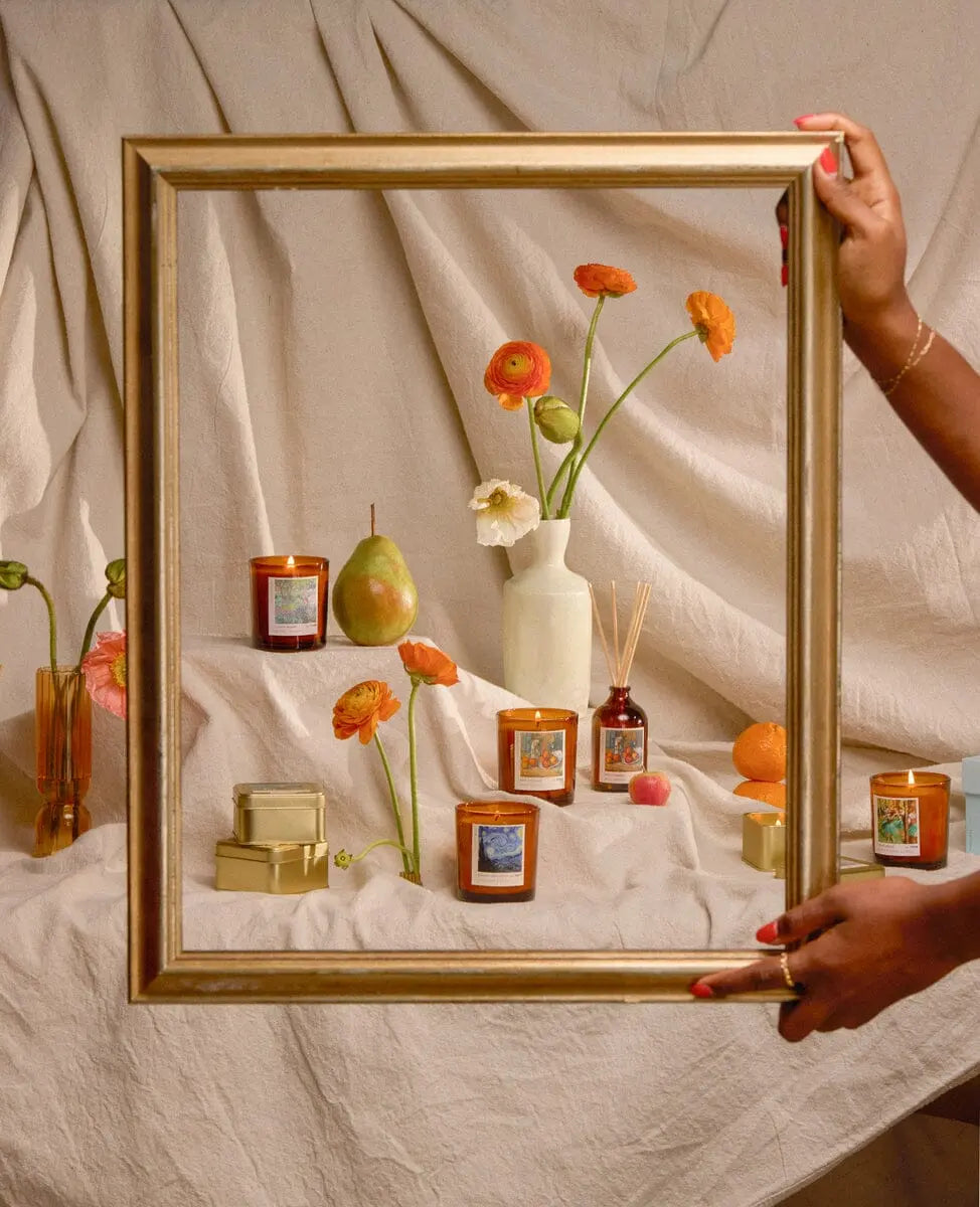A person holds a gold frame before candles, the Impressionist Diffuser - Claude Monet Secret Garden, floral vases, fruits, and small boxes on beige fabric—creating a scene reminiscent of Impressionist art.