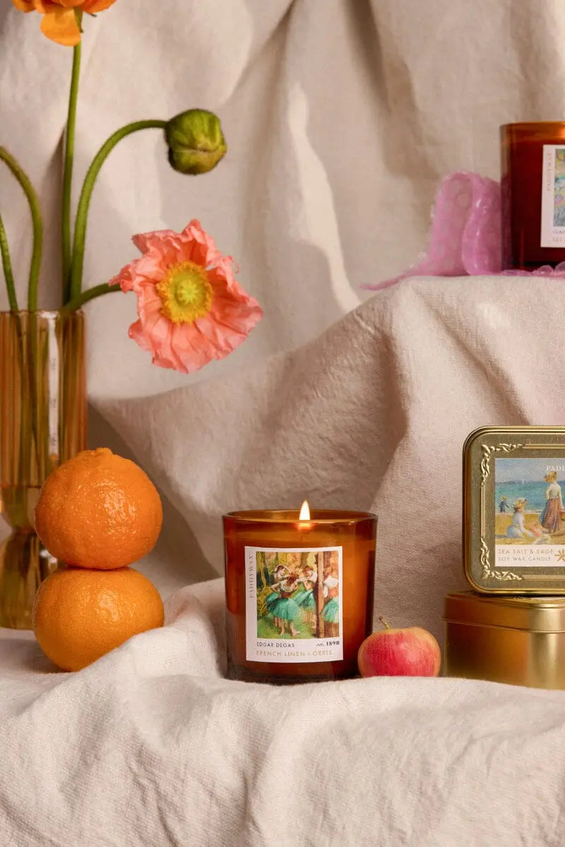The Impressionist Candle - Renoir Sea Salt + Sage, in lit amber glass with a vintage-style label, sits near fruit, a gold tin, and flowers—perfect for lovers of art-inspired fragrances.