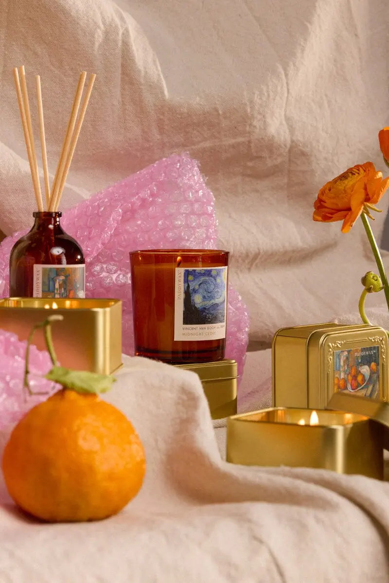 The Impressionist Candle - Vincent Midnight Cedre features glass and tin containers with decorative labels, shown with a reed diffuser bottle, orange, and orange flower on beige fabric against pink bubble wrap.