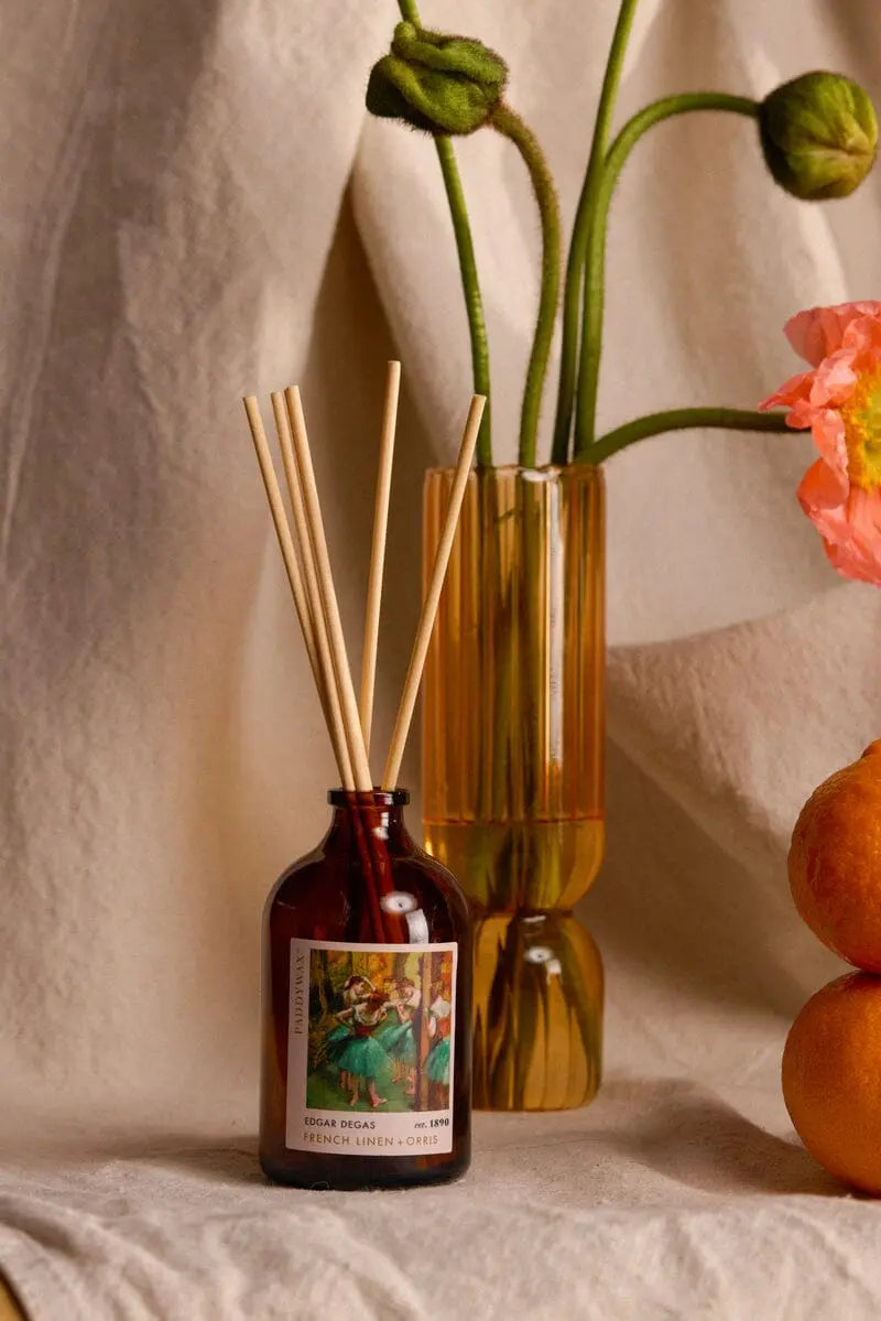 The Impressionist Diffuser - Degas French Linen + Orris sits on a beige cloth near an amber vase with poppies and oranges, evoking artistic fragrance and the essence of Impressionist masterpieces.
