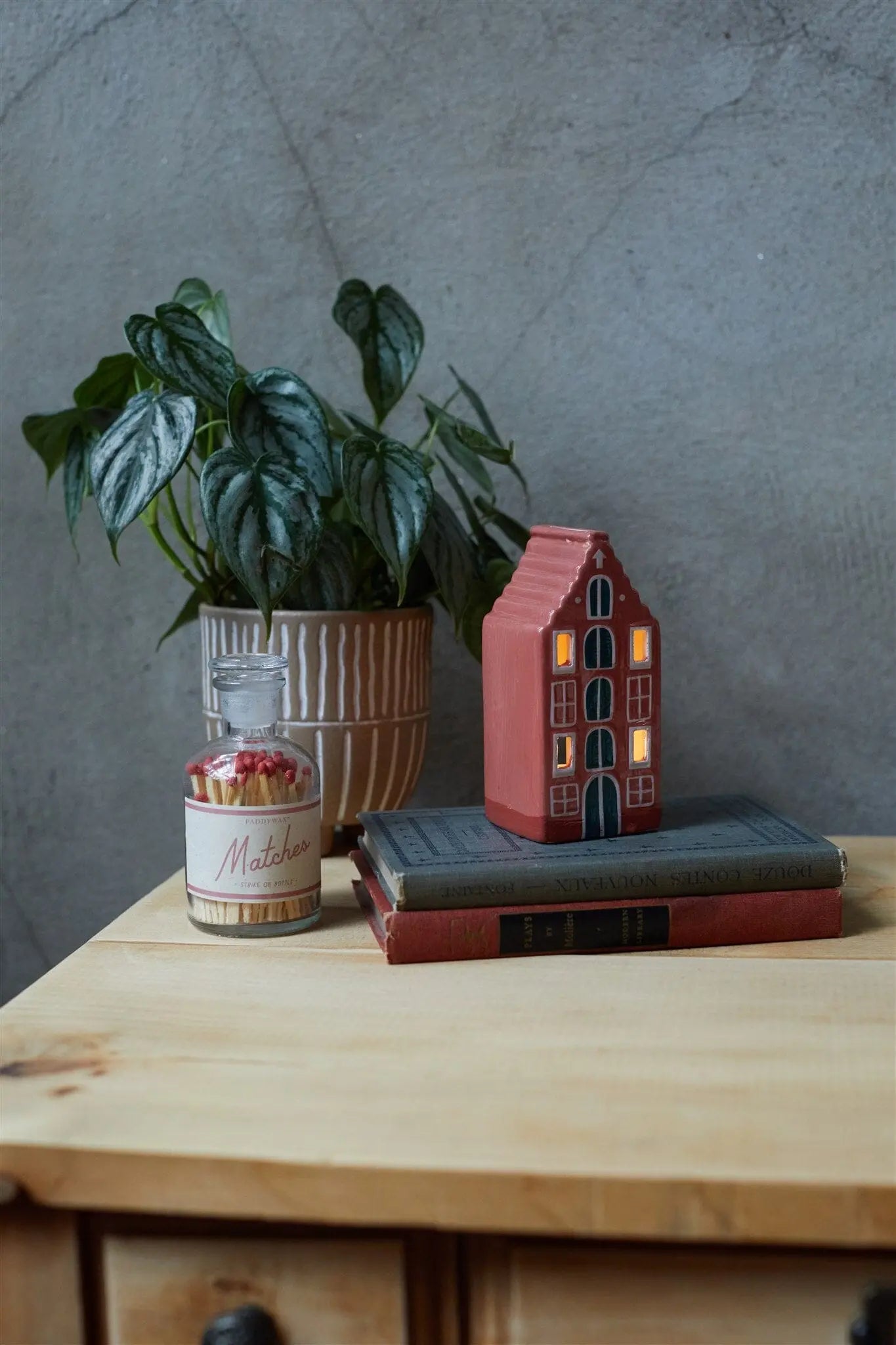 Number 02 of a Collectible Series | The Amsterdam House, a tea light holder shaped like Amsterdam architecture, sits on a wooden table alongside a potted plant, jar of matches, and two stacked books against a gray wall.