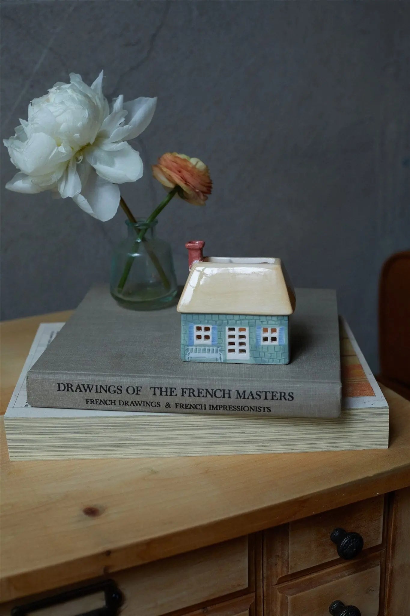 Number 04 of a Collectible Series, The English Cottage—a small ceramic house perfect as a tea light holder or décor—rests on two books beside a glass vase with white and orange flowers atop a wooden table.