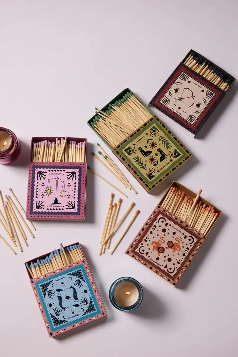 An open Jumbo Boxed Matches - Bow & Arrow set displays its matches scattered on a light surface. Two small lit candles add a cozy touch to the scene.