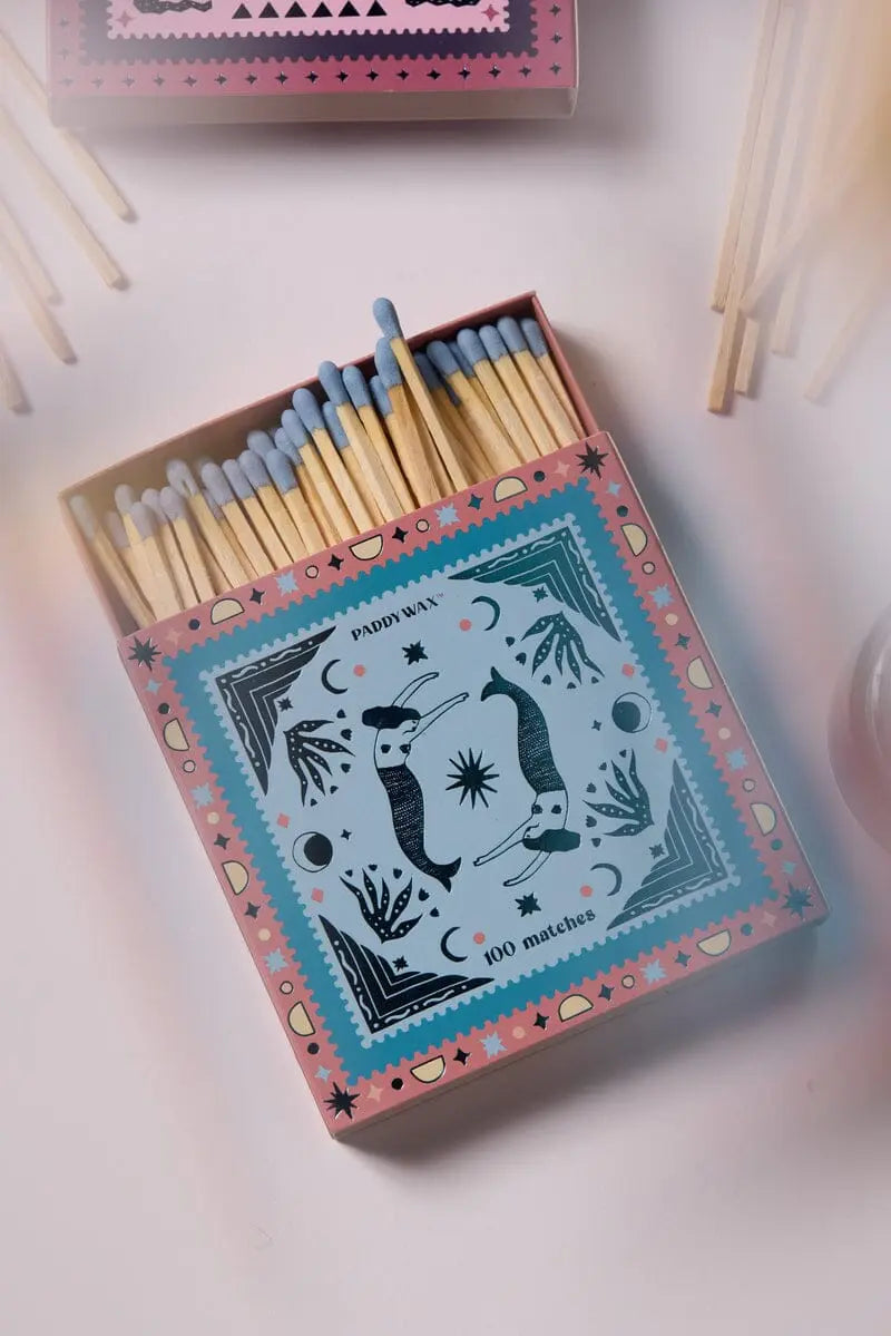 An open Jumbo Boxed Matches - Mermaids, featuring blue-tipped matches and a mermaid-themed illustrated lid, rests on a light surface with some loose matches nearby—perfect for lighting your favorite Paddywax candle.