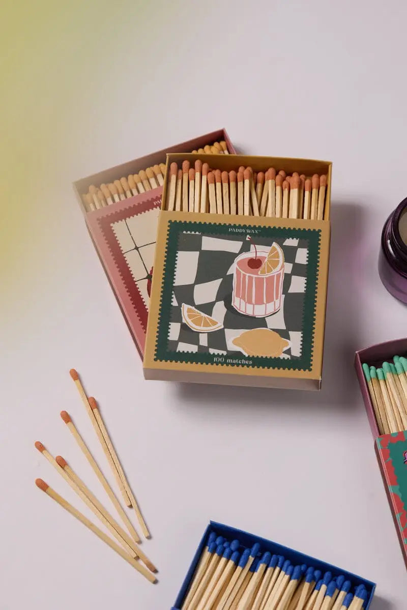 Multiple matchboxes with colorful tips are arranged on a white surface; the central box, the Jumbo Boxed Matches - Cocktail, features a drink, lemon slices, and a checkered background.