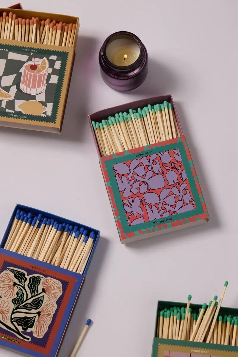 Jumbo Boxed Matches - Abstract Floral are arranged around a lit purple Paddywax candle on a white surface.
