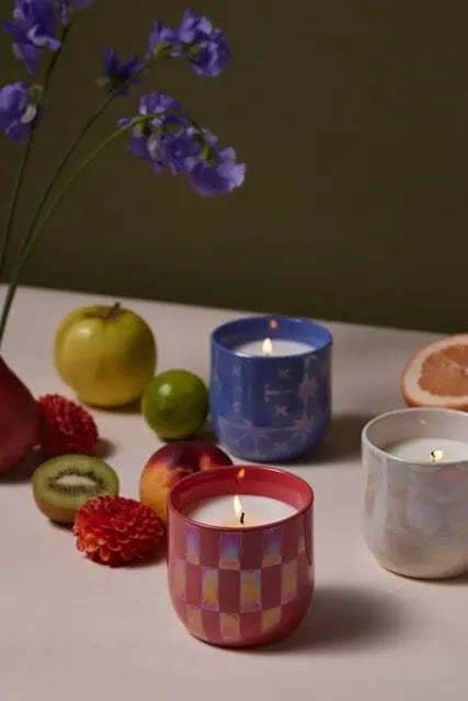 Three lit candles, including the Lustre 10 oz. Candle - Sapphire Waters in an iridescent holder, are arranged on a table with flowers, assorted fruits, and a green backdrop.