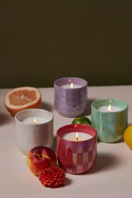 Four lit Lustre 10 oz. Tobacco Vanilla candles in colorful checkered ceramic vessels are arranged on a table with fruit and a red flower. A green background adds to the fresh ambiance.