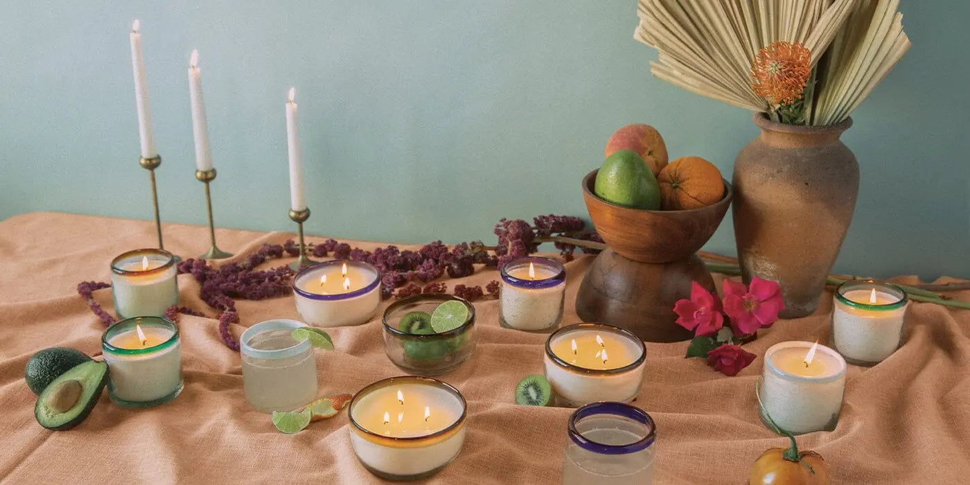 A table display features the La Playa 9 oz Candle - Salted Blue Agave, recycled glass cups, sliced fruits, avocados, flowers, dried palm leaves in a vase, and a wooden fruit bowl—ideal for a recipe blog shoot with a light green backdrop.