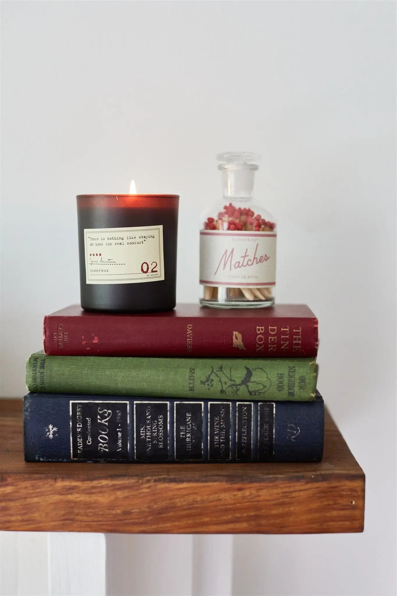 A Library 6 oz Candle - Jane Austen and a glass jar of matches sit atop three stacked hardcover books, creating a cozy space for book lovers on a wooden shelf against a plain white wall.