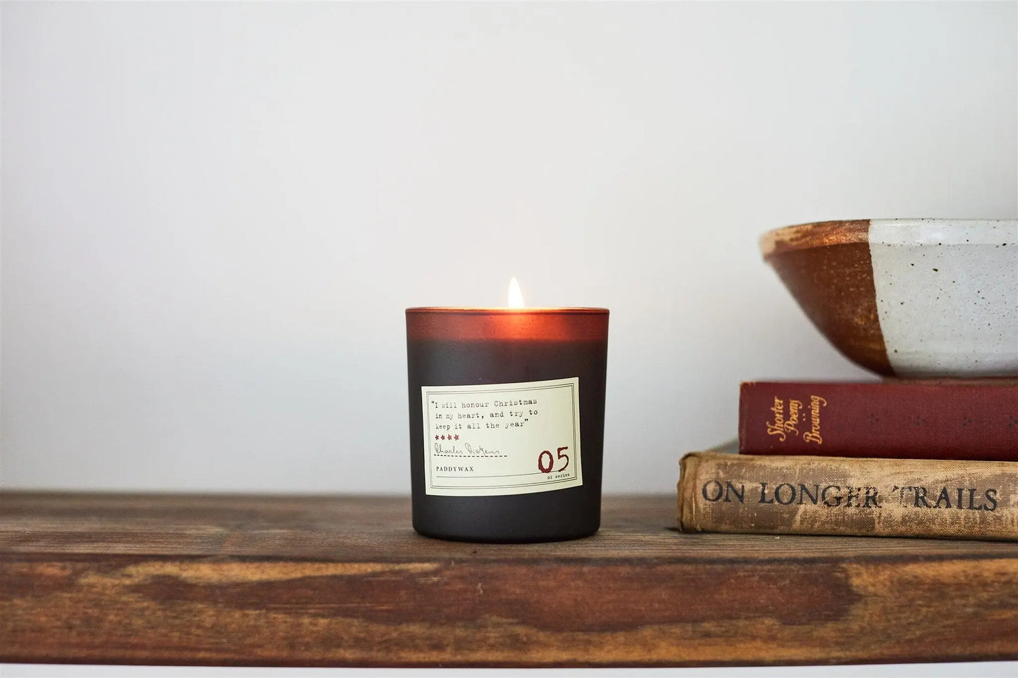A lit Library 6 oz Candle - Charles Dickens in a brown glass jar rests on a wooden shelf beside a ceramic bowl and two stacked books, including ON LONGER TRAILS—ideal for book lovers who cherish literary-inspired scents.