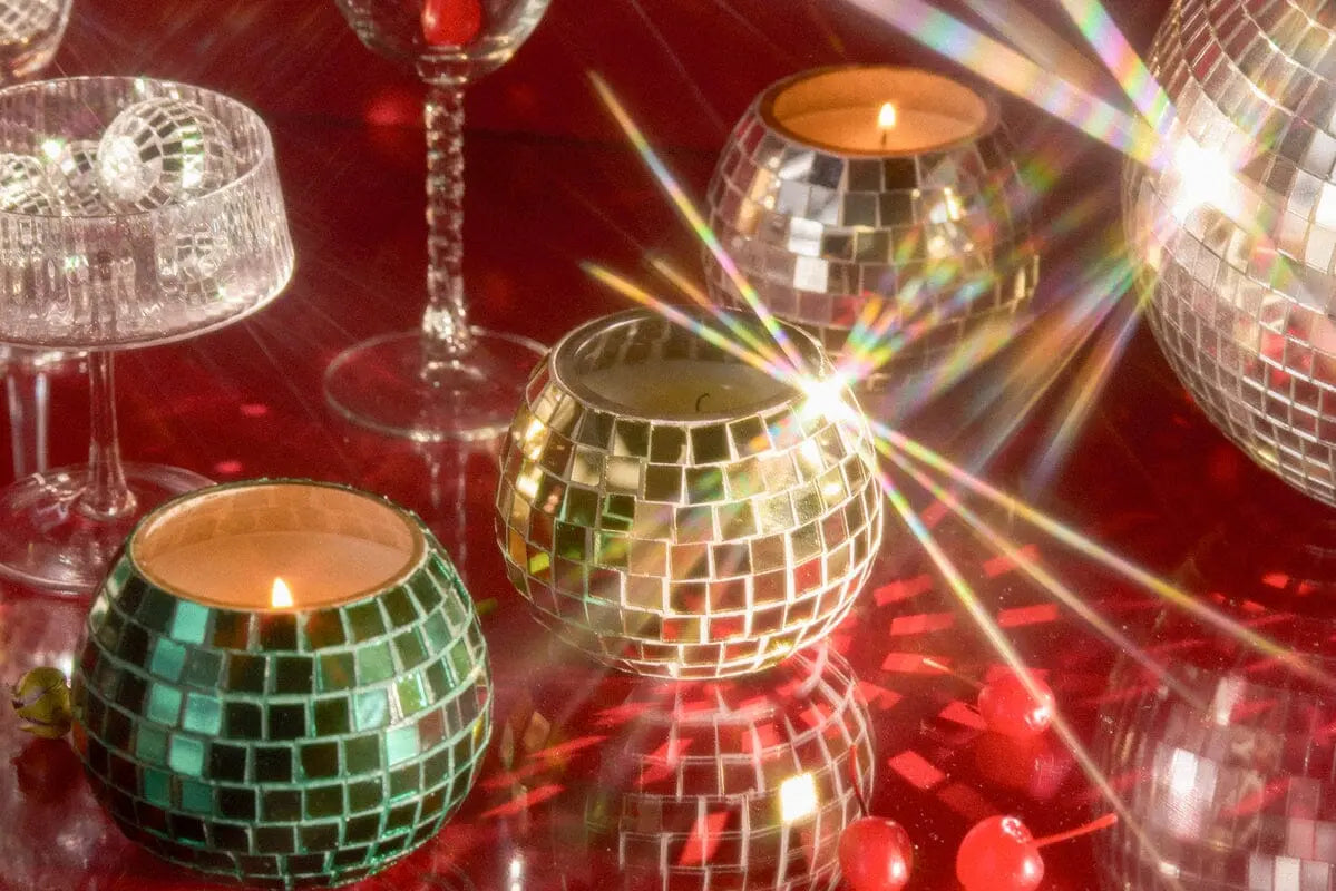 The Magique Green Disco Ball - Tobac & Pine candle casts a warm glow atop a reflective red surface, surrounded by decorative beads and elegant glassware—adding an eye-catching touch to your home decor.