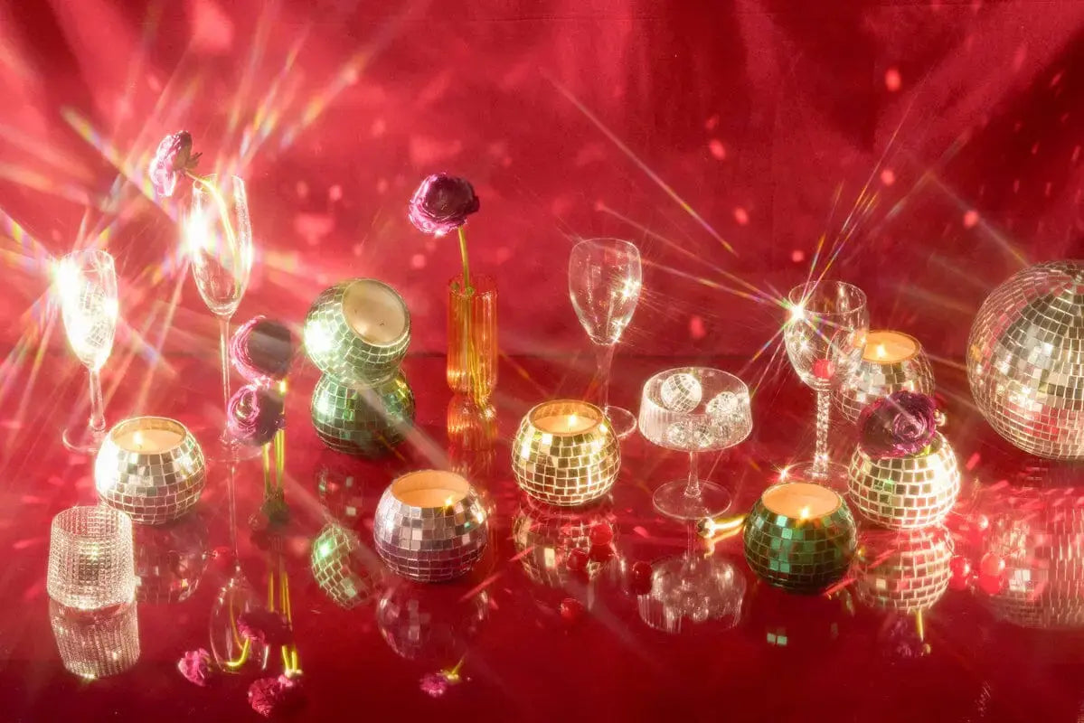 The Magique Green Disco Ball - Lavender Fern is displayed on a reflective red surface, surrounded by glassware and hand-crafted mirror ball candles with sparkling light reflections.