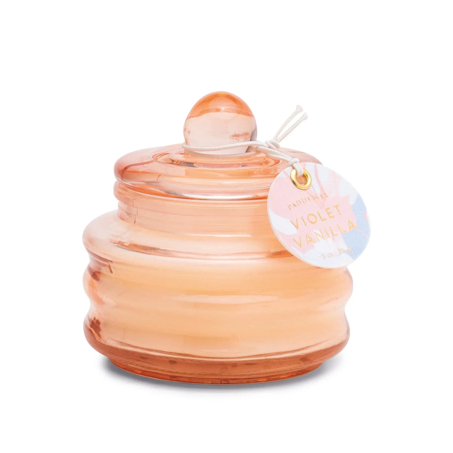 The Beam 3 oz. Candle - Violet Vanilla features a round, peach-colored glass vessel with a matching lid and tag, adding elegance to any space—ideal for small treasures or enjoying a gentle spring scent.