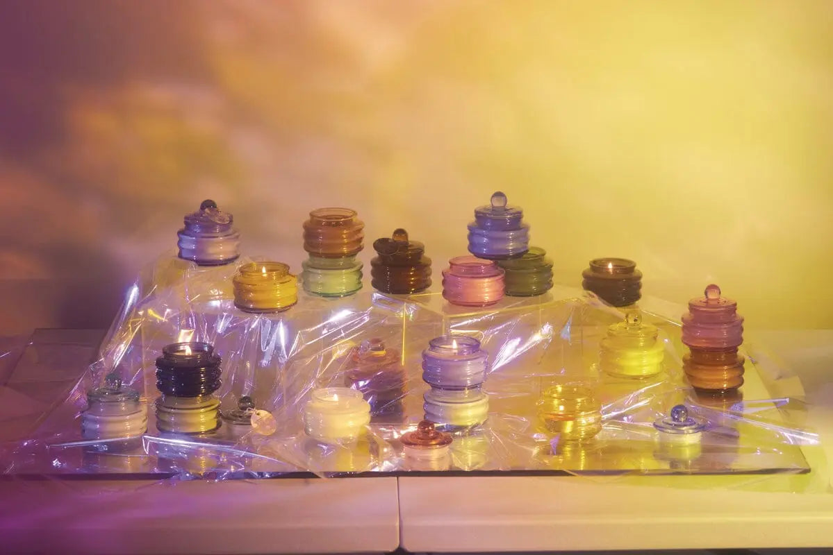 Stacks of vibrant, translucent discs and glass spheres sit on a plastic surface beside the Beam 3oz Glass Candle - Pepper + Plum, all bathed in ambient light with a yellow and purple gradient backdrop.