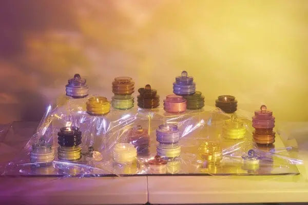 Stacks of Beam 3oz Glass Candle - Pomelo Rose are arranged on a plastic-covered surface, illuminated by soft yellow and purple ambient light for a dreamy atmosphere.