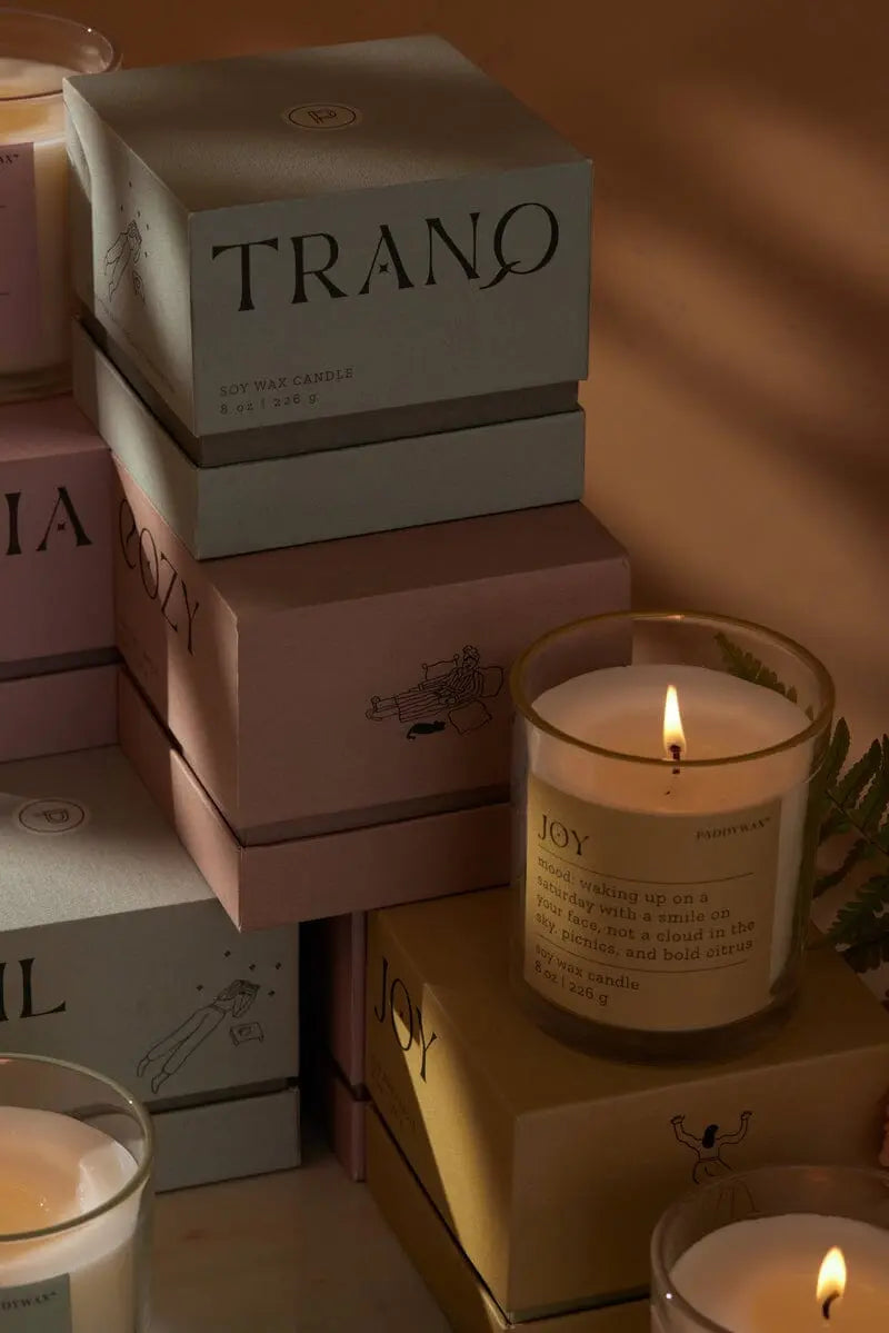 A lit Mood Collection 8oz - Misted Lime Joy candle rests on pastel boxes labeled TRANQ and COZY, radiating an uplifting Joy fragrance with a subtle misted lime aroma in the softly lit background.