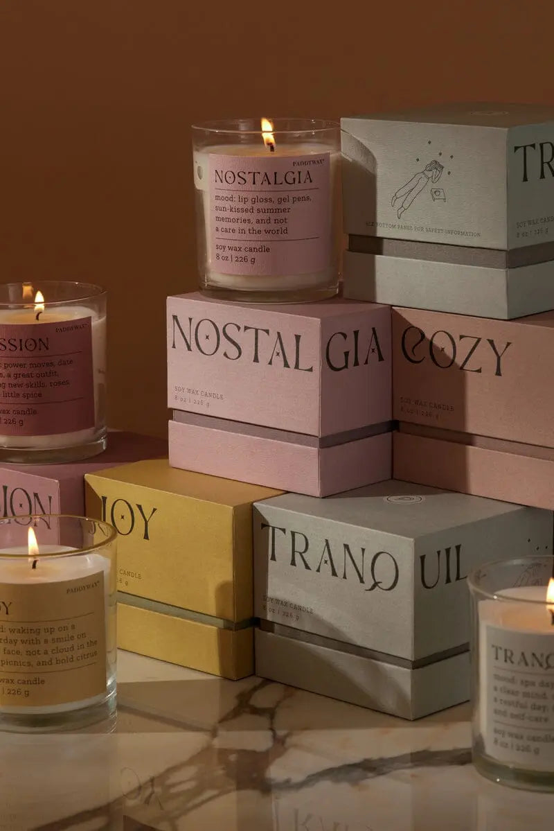 A lit Pepper + Plum Nostalgia Mood Collection 8oz candle in a glass jar is displayed among pastel boxes labeled Nostalgia, Cozy, Tranquil, and Joy on a neutral-toned surface.