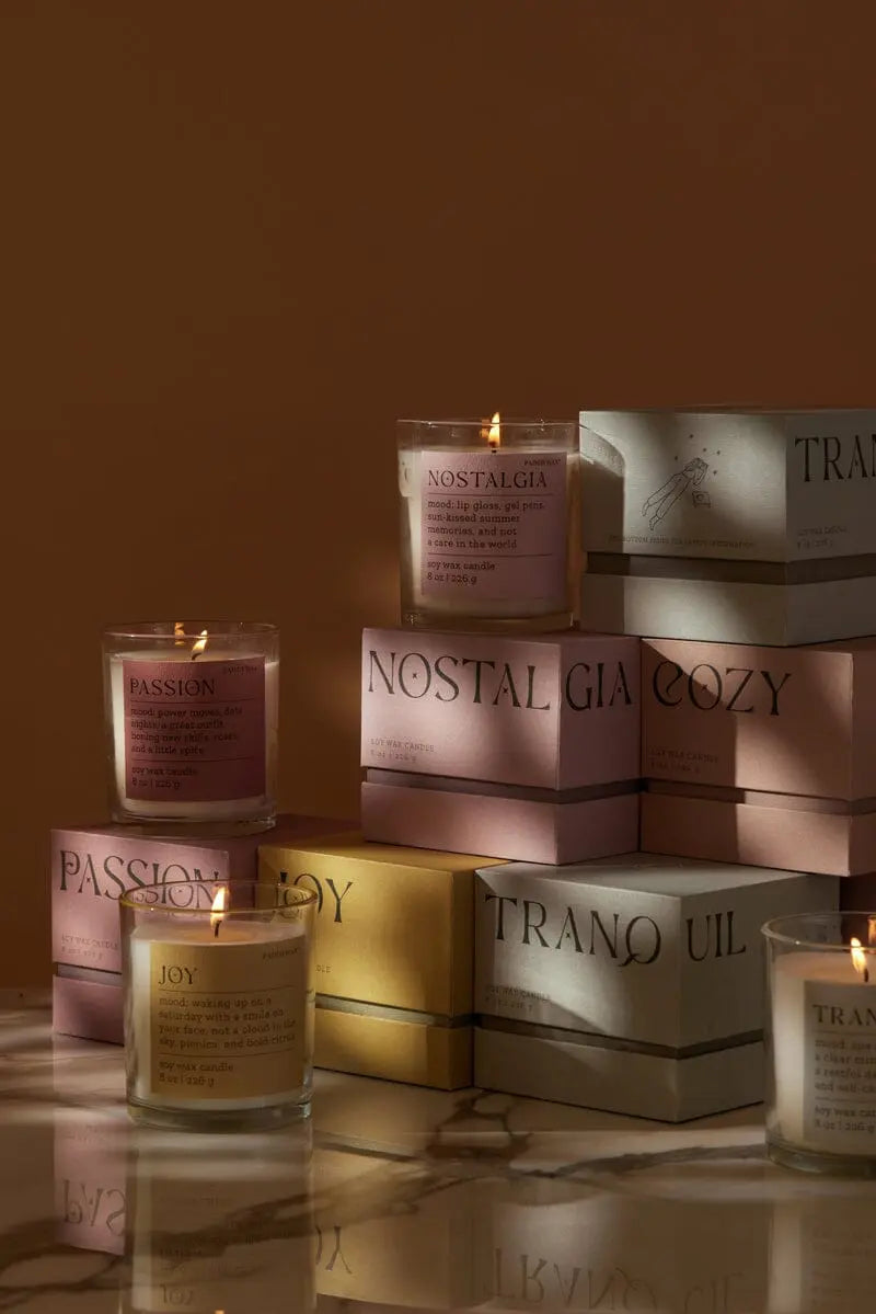 The Saffron Rose "Passion" Mood Collection 8 oz candle, in a glass jar, is arranged with other lit candles on and around pastel boxes labeled Nostalgia, Cozy, Joy, Passion, and Tranquil on a marble surface.