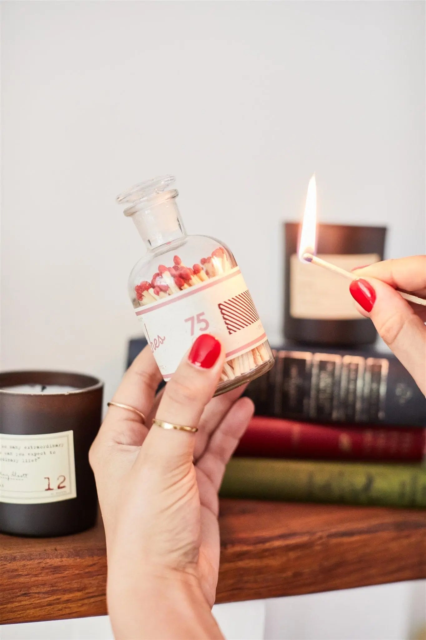 A person with red nail polish lights a wooden match from the Bottle of Matches luxe glass bottle, with books and candles in the background.