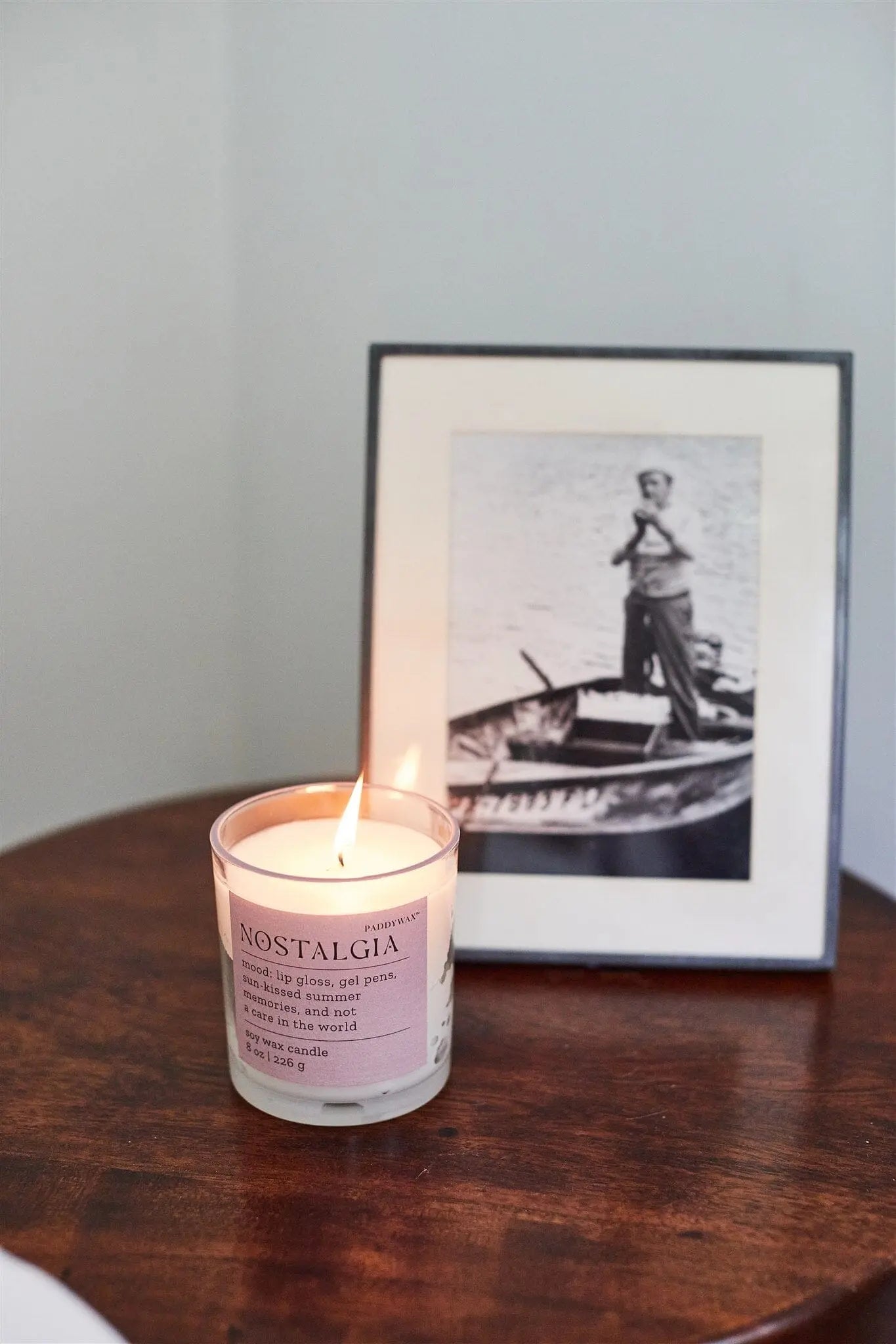 A lit Mood Collection 8oz Pepper + Plum Nostalgia candle rests on a wooden table beside a framed black-and-white photo of someone standing in a small boat.