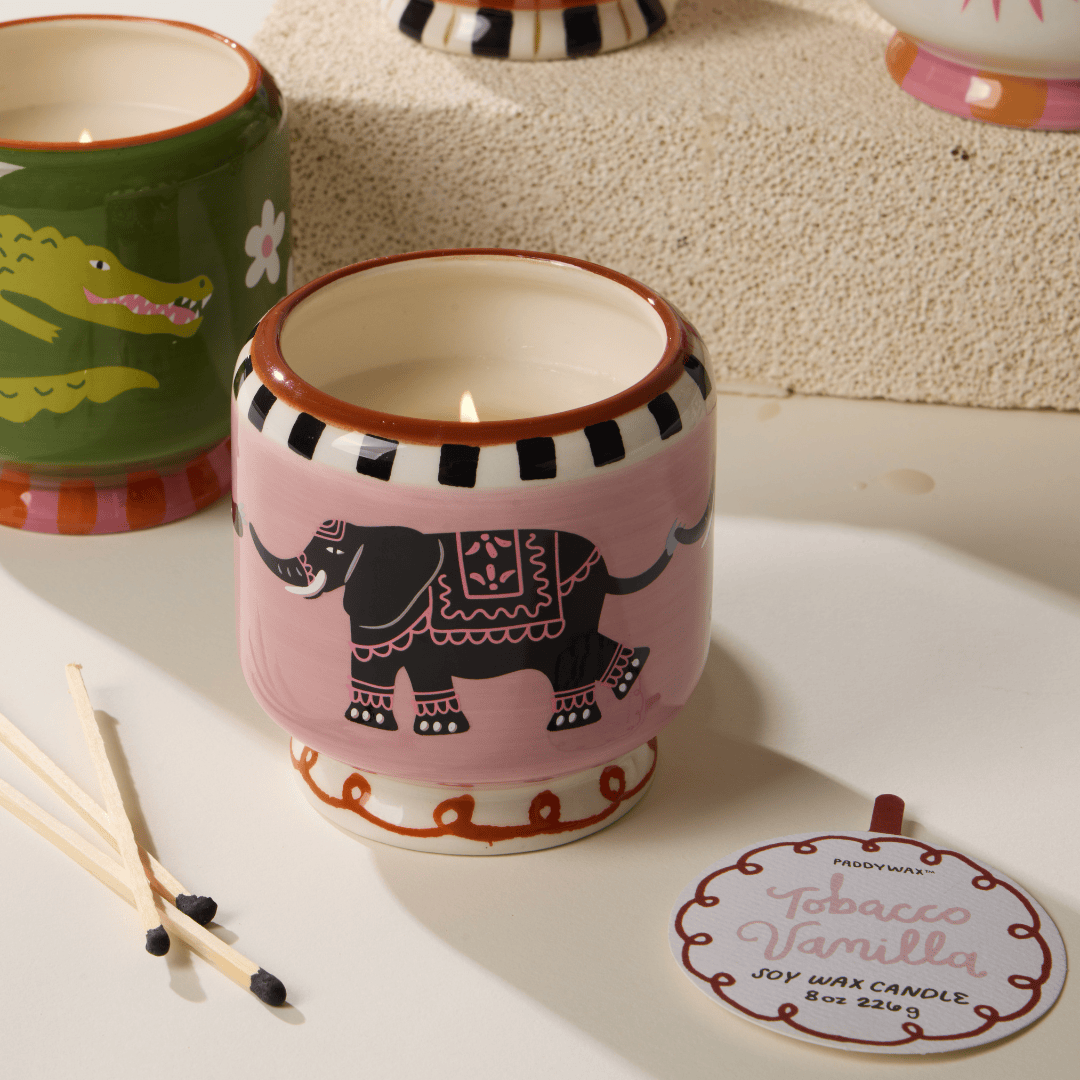 The Tobacco & Vanilla - A Dopo 8 oz Candle features bold fragrances and comes with a ceramic candle holder showcasing a black elephant design on a pink background, accompanied by a round tag and wooden matches—ideal for unique decor lovers.