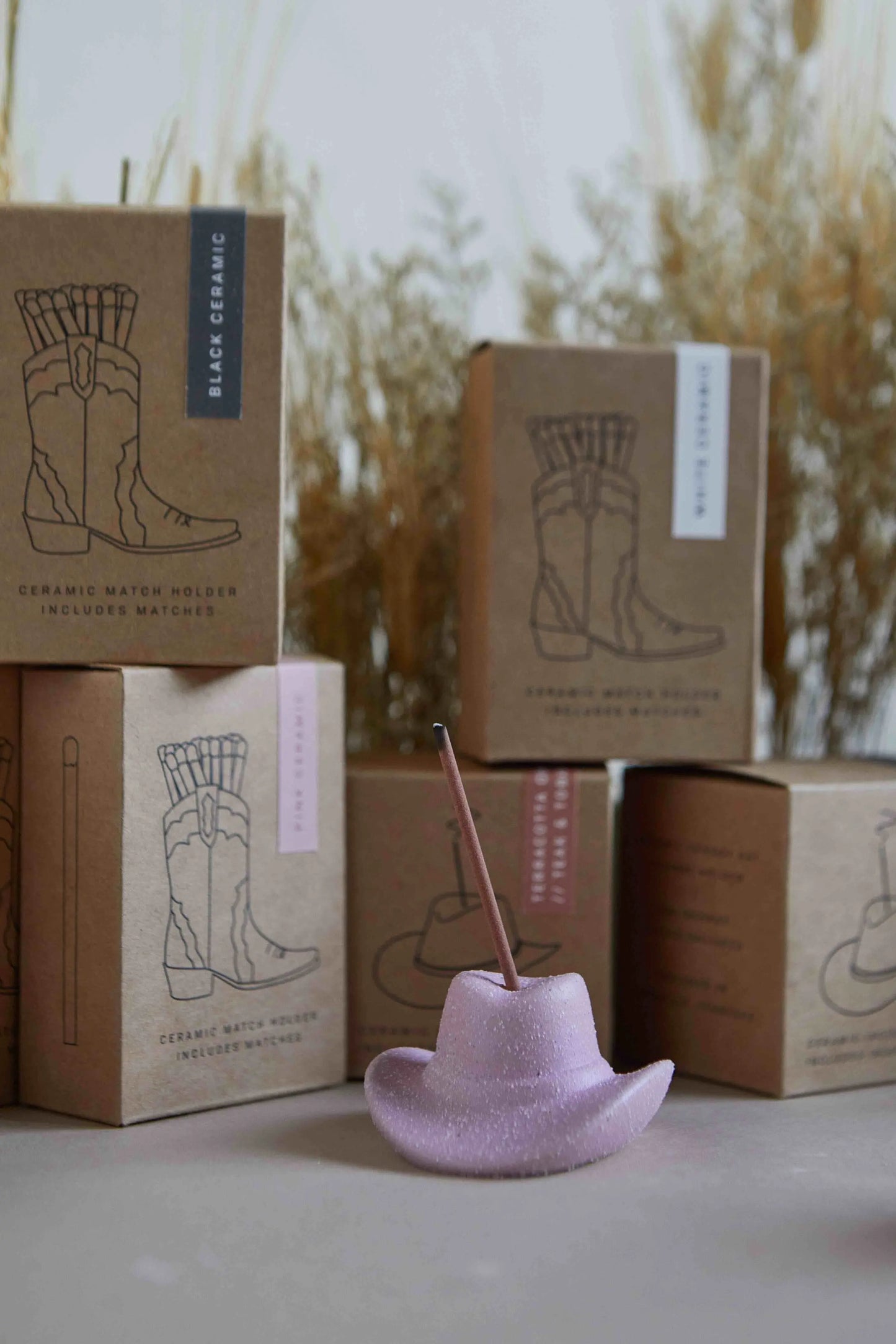 The Cowboy Hat Incense Holder - Pink holds a burning incense stick, with brown boxes featuring cowboy boot designs stacked in the background.