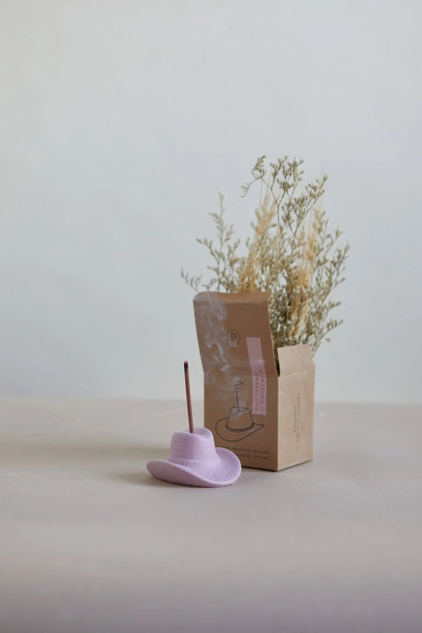 A pink Cowboy Hat Incense Holder cradles a lit stick, smoke rising beside a brown box and dried foliage in the background.