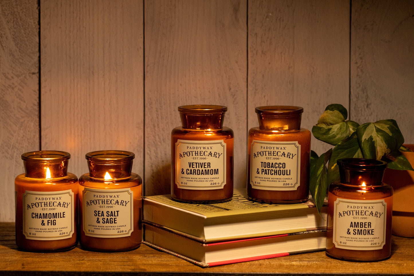 5 of the Apothecary candles, in different fragrances, including Chamomile & Fig. All on a wooden shelf with 2 placed on books