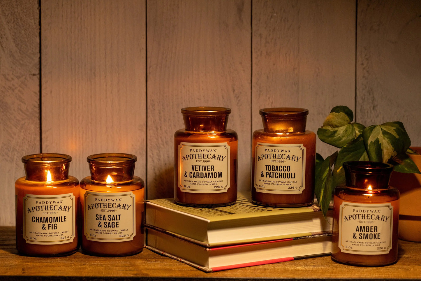 The Apothecary 8 oz Candle - Amber + Smoke, housed in a vintage apothecary-style glass jar, is shown alongside four similar candles on closed books. Two are lit, with a small green plant nearby and white wood planks as the backdrop.