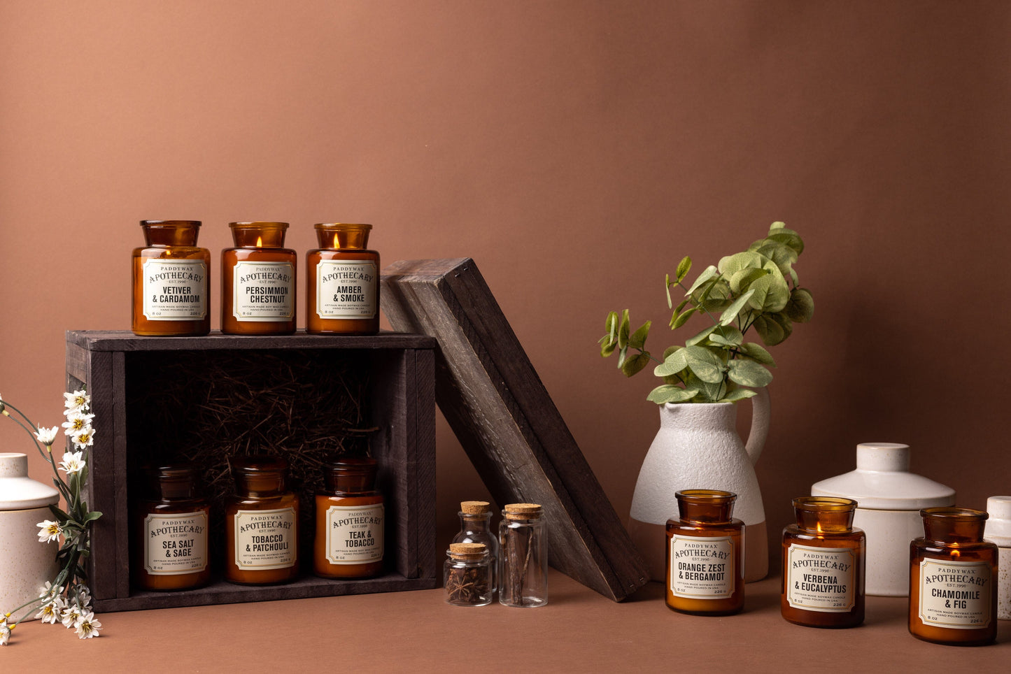 The Apothecary candle collection with 3 on a wooden shelf, 3 in a wooden shelf, and 3 to the left of the wooden shelf with flowers and white jugs