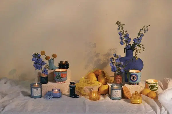 A display featuring the Coastal 7oz Candle - Coconut Husk, blue vases with flowers, lemons, a net bag, sea glass, and small containers arranged on a white cloth against a neutral background.
