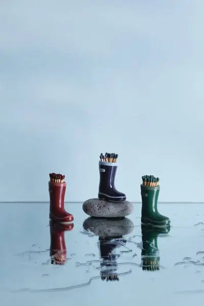 Three dark green Wellington Boot Matches containers, reminiscent of ceramic match holders, are filled with pencils and arranged on a reflective surface, with the middle one set atop a smooth stone.