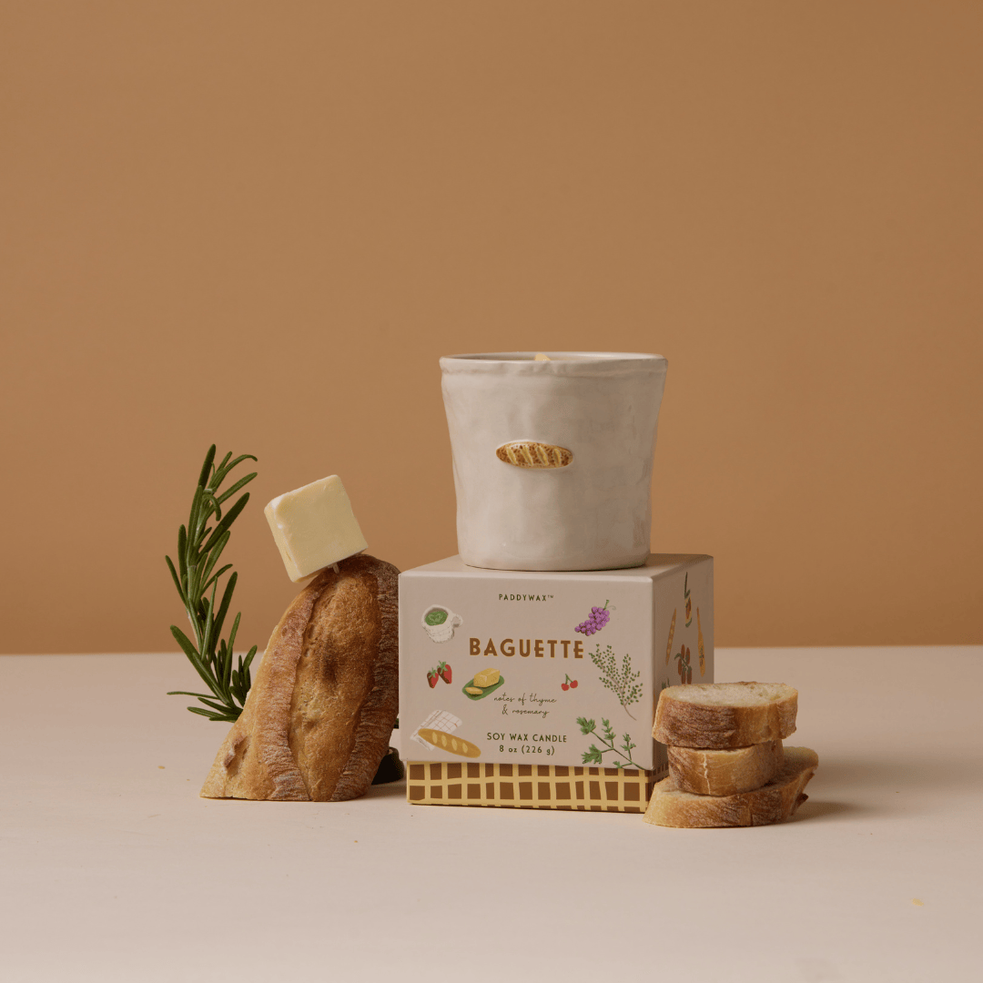 The Baguette - Bistro 8 oz Candle sits atop its box, surrounded by a baguette, rosemary, butter pat, and bread slices—a cozy setting for this aromatic ceramic candle against a beige background.