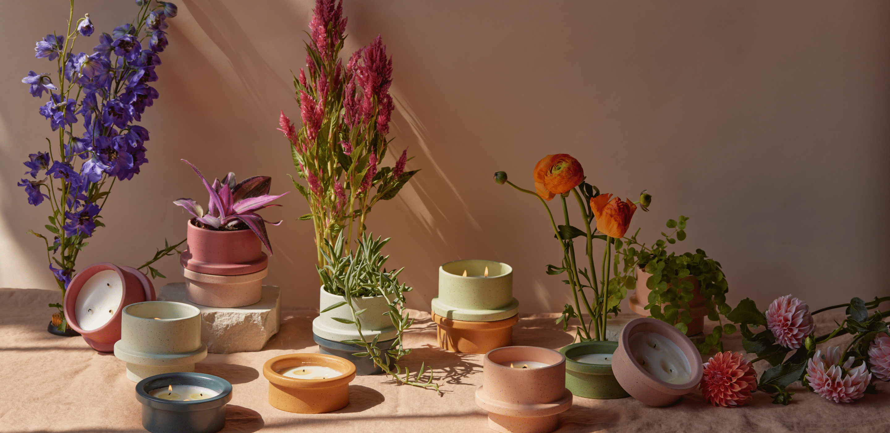 Assorted pastel ceramic candles and pots are arranged on a table with colorful flowers and plants, lit by natural sunlight against a beige background.