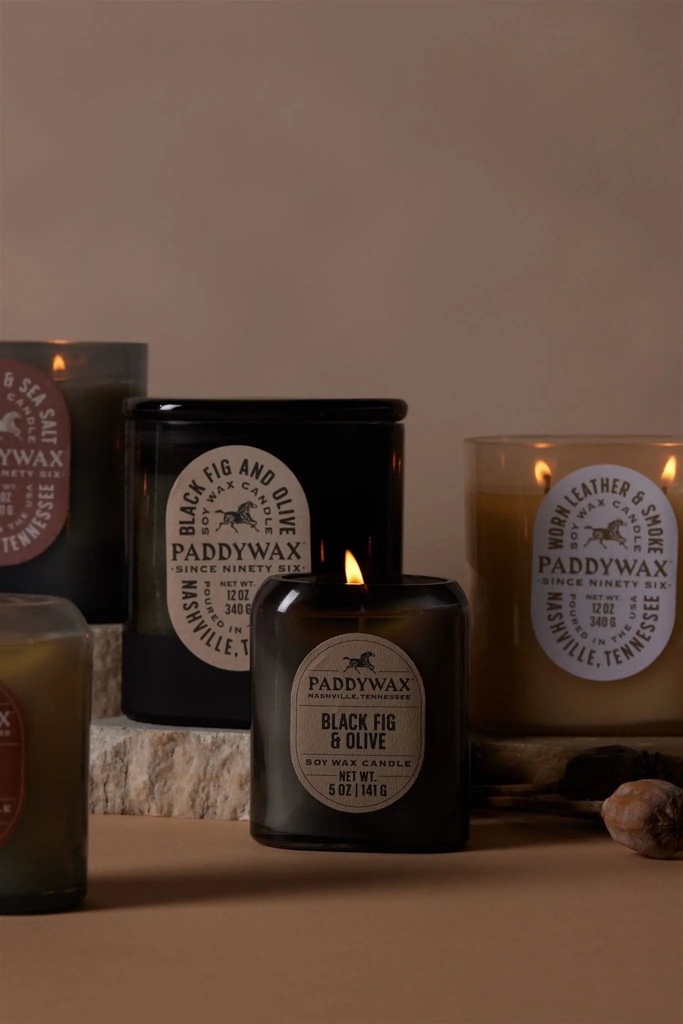 The Vista 5 oz Candle in Black Fig & Olive sits lit among other Paddywax candles on a beige surface, its luxurious scent enhancing any modern home décor.